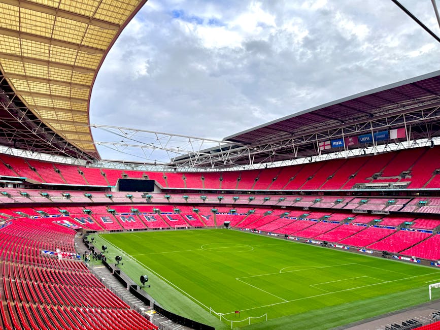 A large football stadium with a partially covered roof structure showing exposed white steel beams and yellow roofing panels overhead. The seating area is filled with bright red plastic chairs arrange